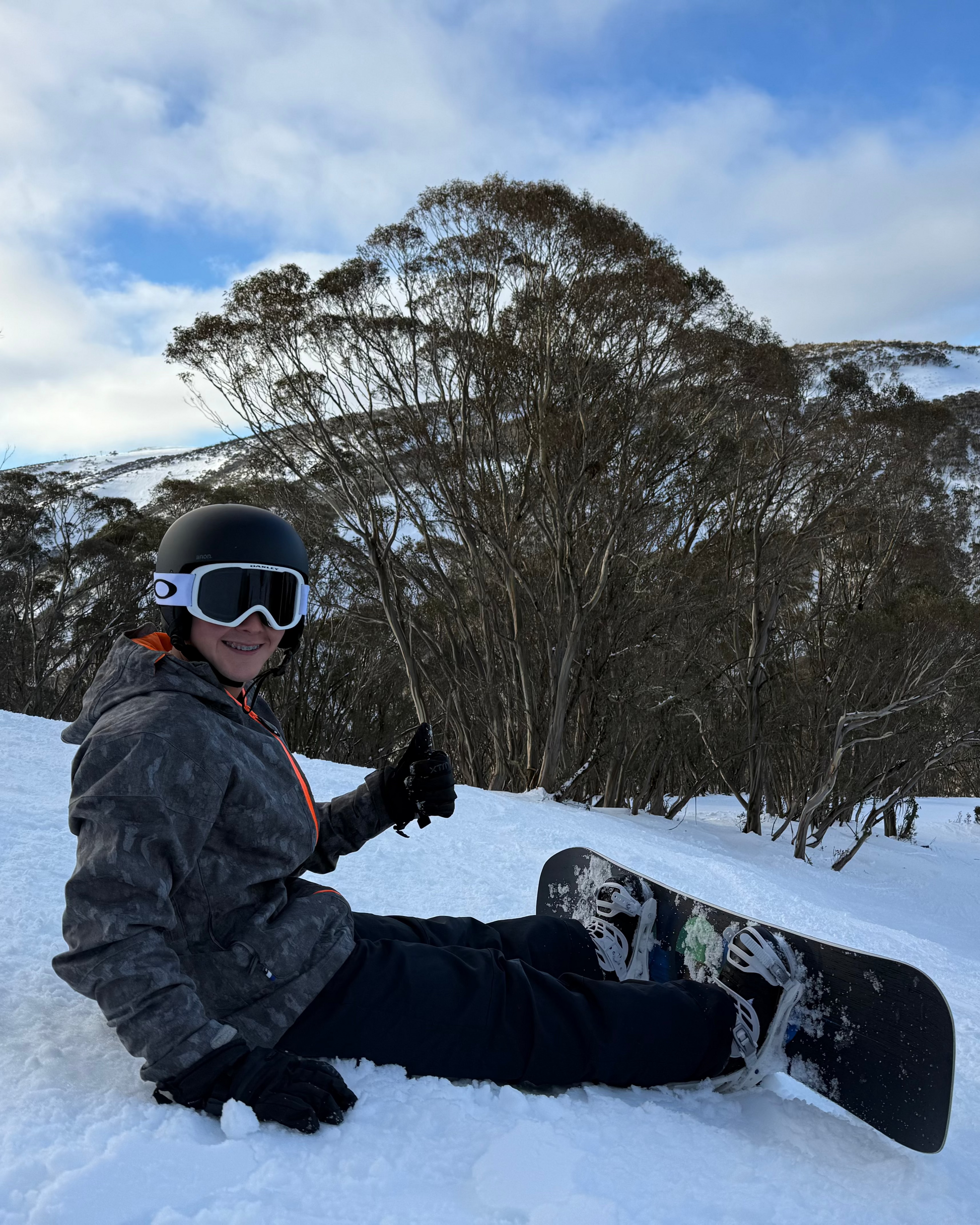 Person sitting on a snow-covered slope with a snowboard, surrounded by trees and mountains.